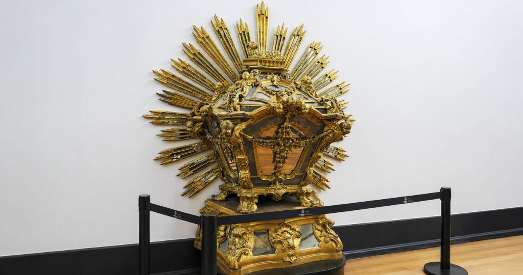 Fotografia colorida e detalhada da Urna do Santíssimo Sacramento, uma peça de talha dourada barroca projetada por Nicolau Nasoni, exibida em um museu. A urna opulenta está centrada contra uma parede branca limpa e repousa sobre uma base dourada correspondente. É uma estrutura complexa, ricamente decorada com esculturas de anjos e ornamentos de folhas. O topo é encimado pelo Cordeiro de Deus reclinado. Atrás da urna, um grande resplendor em forma de sol com raios dourados e espelhados se espalha, criando um efeito visual radiante. Uma fita preta de barreira de segurança, sustentada por postes pretos, está disposta em primeiro plano em um piso de madeira clara e um rodapé preto. A luz é suave e focada na peça.