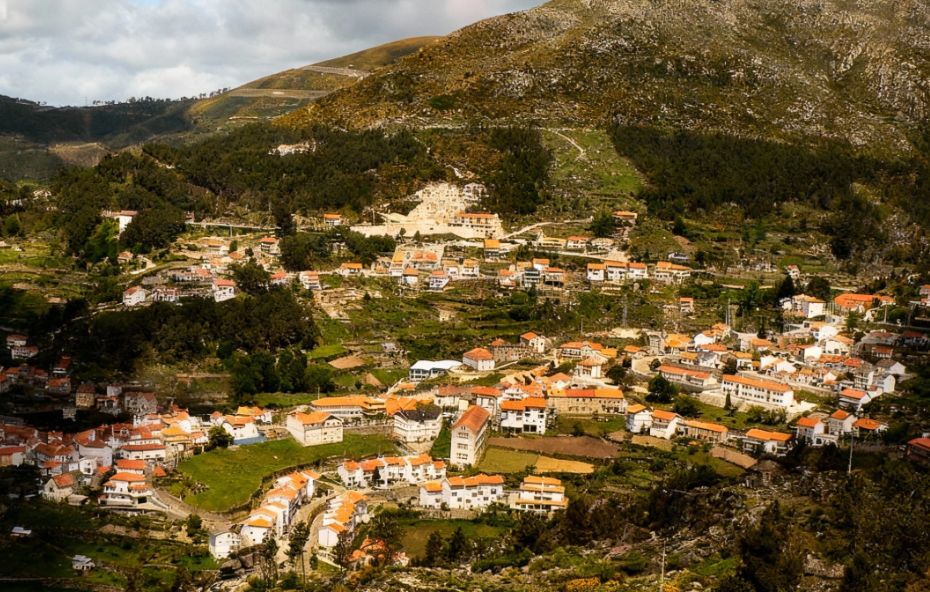Fotografia aérea da aldeia de Loriga, em Portugal, exibindo casas brancas cercadas por montanhas rochosas e encostas verdes esculpidas em socalcos sob a luz do sol.