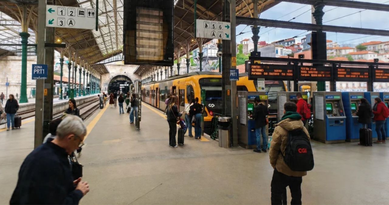 Passageiros utilizam máquinas de bilhetes azuis e amarelas na plataforma da Estação de São Bento com um trem ao fundo.