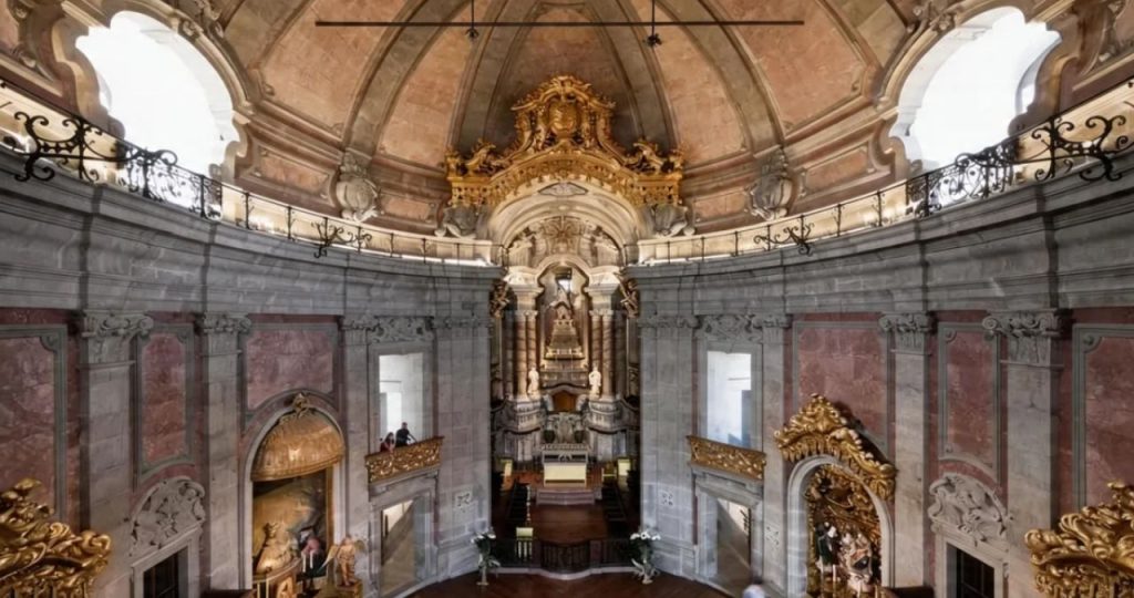 Fotografia em ângulo baixo do interior ornamentado da Igreja dos Clérigos, focando na capela-mor e na cúpula no Porto, Portugal. A imagem captura a arquitetura de pedra cinza robusta da nave que sobe até a cúpula elíptica. A capela-mor no fundo apresenta um retábulo de talha dourada complexo e ricamente esculpido, com esculturas religiosas e ornamentos. Acima do retábulo, uma grande estrutura de coroamento dourada e ornamentada domina, contrastando com o teto de pedra. Galerias e balaustradas de ferro forjado emolduram os níveis superiores, com janelas em arco permitindo a entrada de luz natural. Altares laterais dourados são visíveis nas paredes laterais, e bancos de madeira estão organizados na nave. A luz foca na ação do restauro, realçando os detalhes policromados e a concentração da restauradora.