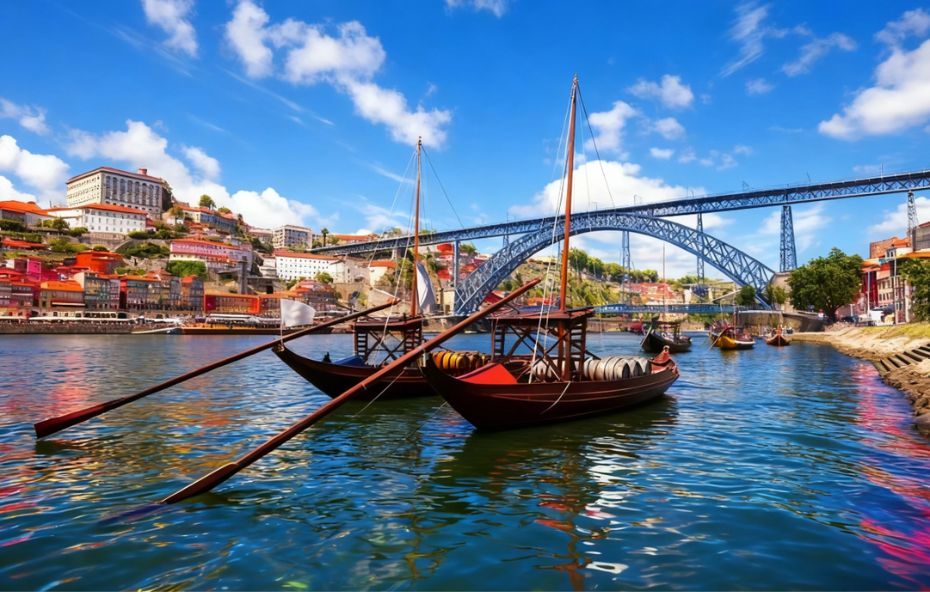 Fotografia colorida do Rio Douro em um dia ensolarado exibindo dois barcos rabelos tradicionais em primeiro plano com a Ponte Dom Luís I ao fundo unindo Porto e Vila Nova de Gaia.