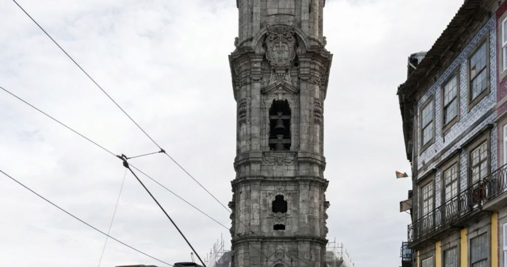 Fotografia em ângulo baixo focada na parte superior da Torre dos Clérigos feita de granito cinza esculpido. A imagem destaca as janelas em arco onde se veem os sinos de bronze e um brasão ornamentado em relevo logo acima da abertura principal. À direita da torre surge a fachada de um edifício tradicional português com janelas de molduras brancas e azulejos azuis e brancos típicos. No canto inferior esquerdo são visíveis os cabos elétricos aéreos que atravessam a rua. O céu ao fundo está nublado e esbranquiçado criando um contraste suave com a textura rugosa e histórica da pedra monumental do monumento no Porto.