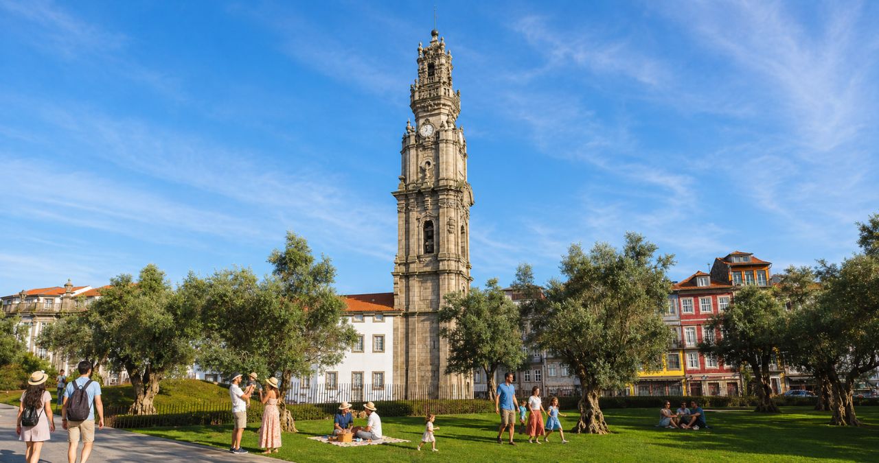 Fotografia colorida e vibrante de um parque gramado em um dia ensolarado no Porto, Portugal. A icônica Torre dos Clérigos de granito, com seus detalhes barrocos e relógio, domina o centro do fundo. No gramado verde, várias pessoas aproveitam o dia: algumas fazem piquenique, outras passeiam com as famílias e um casal caminha por um caminho de pedra em primeiro plano à esquerda. Oliveiras frondosas estão espalhadas pelo jardim e edifícios históricos coloridos alinham a rua ao fundo sob um céu azul claro.