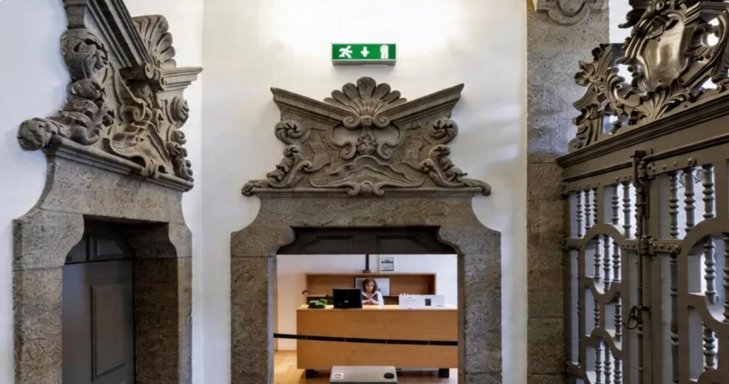 Uma fotografia de ângulo médio em plano geral que captura a entrada principal e recepção da Torre dos Clérigos no Porto, Portugal. A imagem mostra uma imponente e complexa porta de pedra esculpida em estilo barroco e rococó, com um frontão ornamentado que inclui brasões, molduras e detalhes florais. Abaixo da pedra, um portal de recepção moderno com uma estrutura de madeira clara e um balcão de vidro está visível, com uma recepcionista sentada atrás de um computador e uma placa informativa. À esquerda e à direita, outros portais de pedra e uma cerca de madeira e metal com detalhes ornamentais emolduram a entrada. Acima do frontão de pedra, há um letreiro retangular de madeira com o texto em Português: 'Visita ao Coração dos Clériigos: Guia e Acesso'. O chão é de madeira clara. A luz é suave e bem distribuída, destacando as texturas contrastantes da pedra antiga e da madeira moderna.