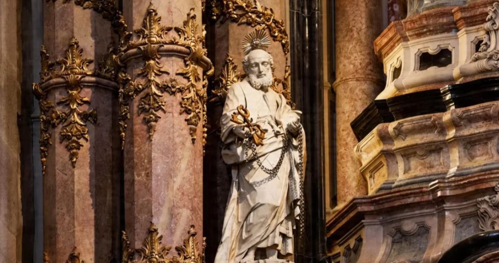 Fotografia em close focada em uma estátua de mármore branco de São Pedro localizada no interior da Igreja dos Clérigos no Porto. A escultura detalhada mostra o santo segurando as chaves do céu com uma das mãos enquanto uma corrente de metal pende de seus braços. Ele possui uma auréola dourada em formato de resplendor sobre a cabeça. Ao lado da estátua há uma imponente coluna de mármore rosado adornada com complexos entalhes de talha dourada em estilo barroco. Uma placa dourada fixada na coluna contém o texto: 'São Pedro: Porteiro dos Céus'. O fundo apresenta outras colunas e elementos arquitetônicos em tons de bege e marrom com acabamentos em mármore escuro criando uma atmosfera solene e histórica.