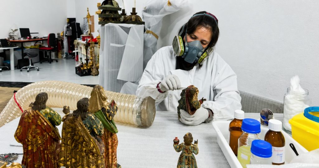 Fotografia em plano médio focada em uma especialista em conservação e restauro em um ateliê de arte. A mulher, vestida com um macacão de proteção branco, luvas de nitrilo e uma máscara respiratória de dupla filtragem, está sentada à frente de uma mesa de trabalho limpa. Ela concentra-se em uma pequena escultura religiosa antiga de madeira policromada que segura com a mão esquerda, enquanto usa uma ferramenta de precisão de metal fina com a direita para realizar um restauro meticuloso em detalhes dourados e verdes. À sua esquerda, sobre um material protetor branco, estão outras esculturas religiosas maiores e douradas, incluindo figuras de santos e a Virgem Maria. Em primeiro plano à direita, há frascos de vidro marrom com solventes e soluções químicas, potes com algodão e uma bandeja de ferramentas organizadas. Ao fundo, o ateliê exibe estantes com materiais, outras peças de arte em processo e um duto de ventilação flexível. A luz foca na ação do restauro, realçando os detalhes policromados e a concentração da restauradora.