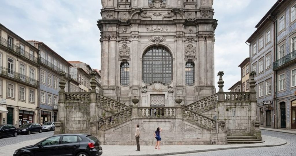 Fotografia frontal da fachada em granito da Igreja dos Clérigos no Porto. Em primeiro plano há uma escadaria monumental dupla com balaustradas de pedra que convergem para o centro da entrada. Duas pessoas estão na calçada em frente ao monumento olhando para cima. A fachada apresenta janelas altas com grades e esculturas barrocas detalhadas em relevo. Edifícios históricos com varandas de ferro e azulejos emolduram a rua em ambos os lados. Um carro preto está estacionado à esquerda e o chão é de paralelepípedos típicos portugueses sob um céu nublado.