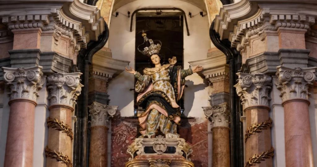 Fotografia centralizada em plano médio da imagem de Nossa Senhora da Assunção no altar-mor da Igreja dos Clérigos no Porto. A escultura policromada veste mantos em tons de dourado, azul e rosa com padrões florais e usa uma coroa prateada ornamentada. Ela está com os braços abertos em um nicho de mármore rosado e bege. O nicho é emoldurado por colunas coríntias de mármore com capitéis detalhados e guirlandas de talha dourada. Uma placa dourada na base da estátua contém o texto: 'Nossa Senhora da Assunção: Padroeira do Templo'. A arquitetura superior apresenta molduras curvas e robustas em pedra cinza que criam uma profundidade dramática sob a iluminação focada.