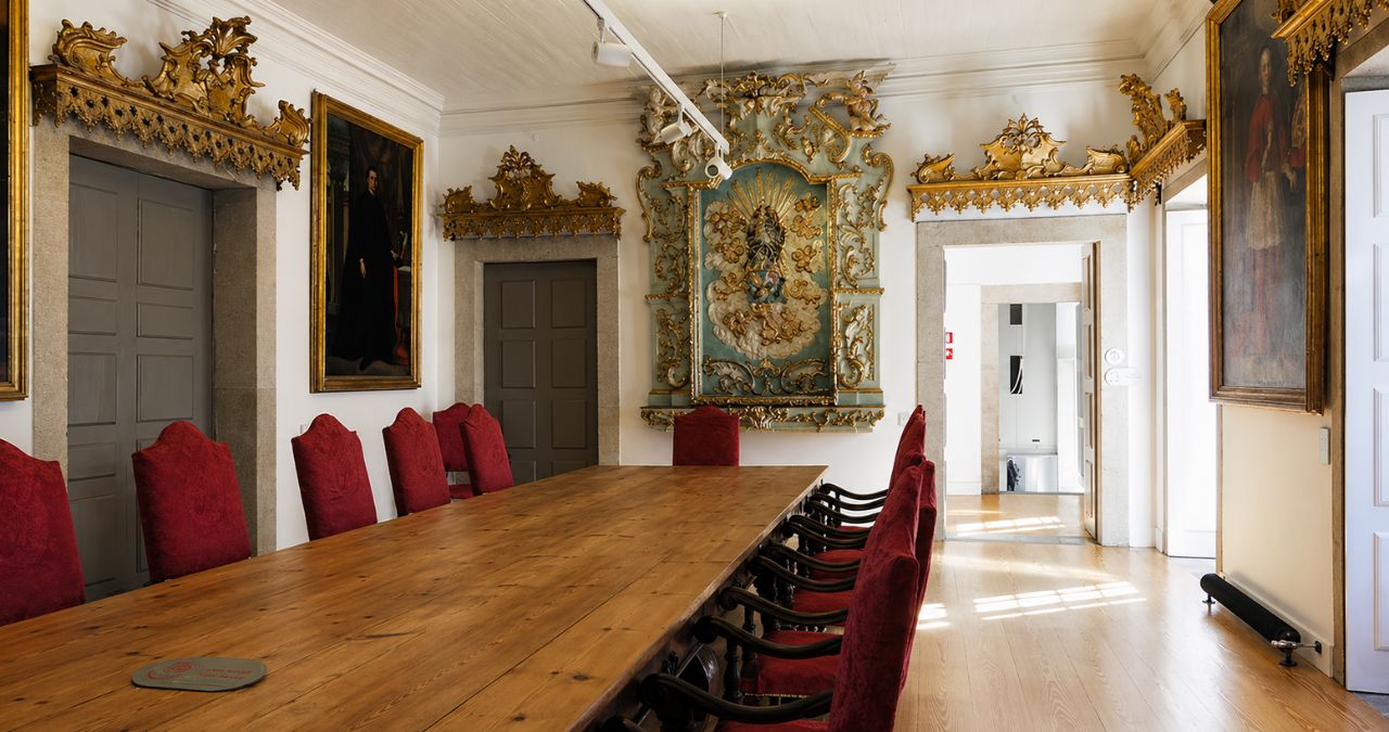 Fotografia colorida e rica do Salão Nobre, uma sala de reuniões histórica na Casa da Irmandade do complexo dos Clérigos no Porto, Portugal. No centro, uma longa mesa de conferências em madeira rústica estende-se, ladeada por várias cadeiras com estofos vermelhos vibrantes e costas altas. As paredes brancas são decoradas com molduras douradas e ornamentadas em estilo barroco e rococó, que se estendem sobre portas de madeira cinzentas e portas abertas. O ponto focal na parede do fundo é um retábulo dourado e turquesa incrivelmente ornamentado, ricamente esculpido, e acima dele há um brasão em cores e detalhes dourados. Pinturas em molduras douradas pesadas, retratando figuras religiosas e históricas, estão penduradas nas paredes lateral e traseira. O chão é feito de tábuas de madeira claras e bem polidas, e a luz solar suave entra pelas janelas e portas abertas, iluminando a sala. A arquitetura é refinada e histórica.