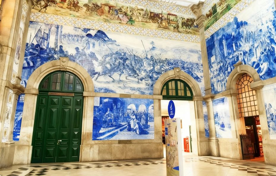 Vista ampla do átrio da Estação de Trem São Bento no Porto mostrando paredes monumentais cobertas por azulejos azuis e brancos com cenas de batalhas e eventos históricos portugueses além de um friso colorido superior ilustrando carruagens antigas e portas verdes clássicas sob arcos de pedra.