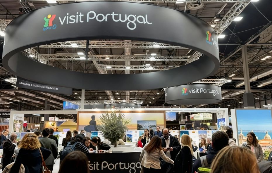 Fotografia do interior do pavilhão na FITUR com grandes estruturas circulares pretas suspensas exibindo o logótipo Visit Portugal. No centro vê-se um balcão de atendimento com pessoas e uma oliveira decorativa.