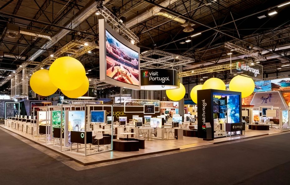 Fotografia de um stand de turismo moderno e iluminado em uma feira de negócios com grandes esferas amarelas suspensas e painéis digitais exibindo imagens de cidades históricas e a marca Visit Portugal.