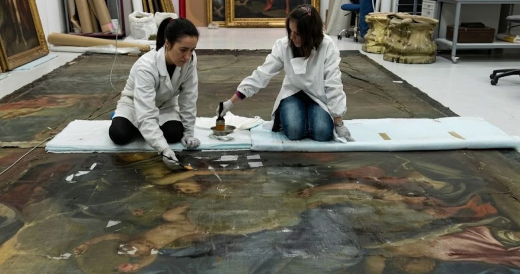 Fotografia em plano aberto capturando duas especialistas em conservação e restauro trabalhando ajoelhadas sobre uma pintura a óleo de grande formato estendida no chão de um ateliê profissional. Ambas vestem jalecos brancos e luvas de proteção enquanto realizam procedimentos técnicos delicados na superfície da obra que apresenta figuras angelicais e motivos clássicos. A restauradora à esquerda utiliza um pincel fino para aplicar solventes ou pigmentos em áreas específicas enquanto a colega à direita manipula um recipiente metálico e ferramentas de limpeza sobre protetores acolchoados brancos. O ambiente do ateliê é amplo com outras molduras douradas e telas ao fundo indicando um processo rigoroso de recuperação patrimonial sob iluminação artificial controlada.