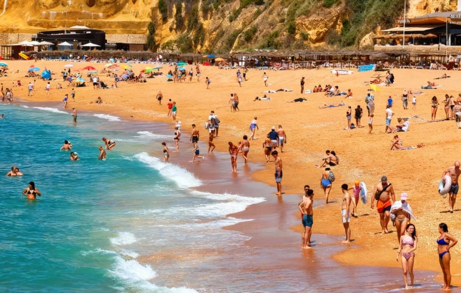 Fotografia panorâmica de uma praia em Portugal com areia dourada repleta de turistas, aproveitando o mar azul e o sol em um dia claro.
