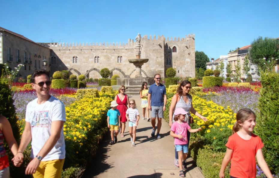 Grupo de crianças e adultos caminhando por um caminho de areia no Jardim de Santa Bárbara em Braga com canteiros geométricos coloridos e muralha medieval ao fundo sob céu azul.