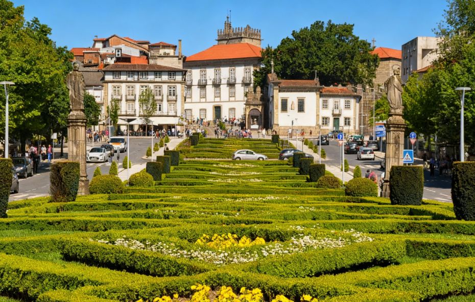 Fotografia colorida mostrando um jardim geométrico com arbustos verdes aparados em labirinto e flores amarelas em primeiro plano. Ao fundo há uma igreja branca clássica e edifícios históricos sob um céu azul claro.