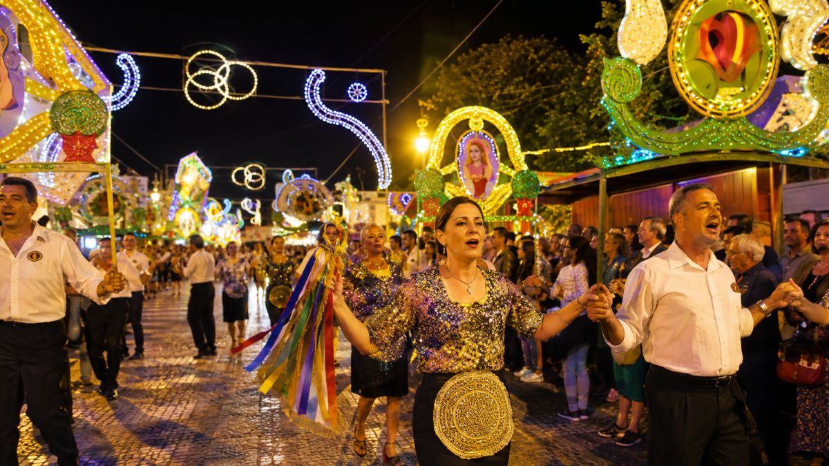 Pessoas desfilam animadas em uma rua iluminada à noite vestindo trajes festivos e brilhantes durante uma celebração tradicional portuguesa.