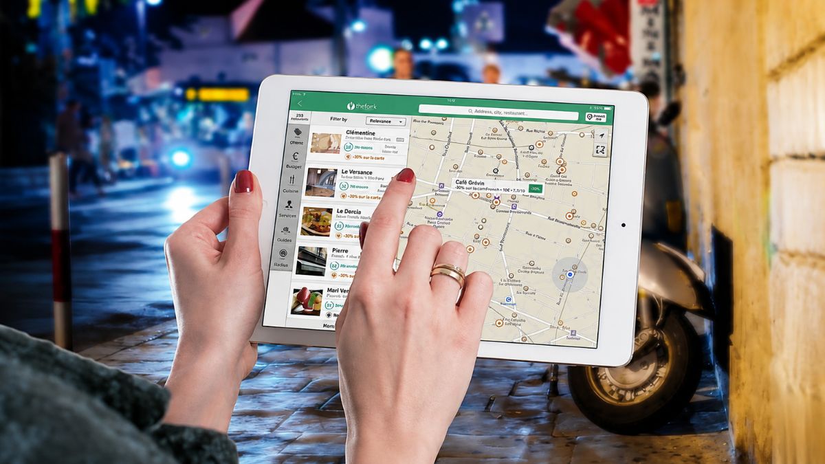 Pessoa segurando um tablet branco e tocando na tela com mapa interativo para buscar localizações em uma rua iluminada à noite.