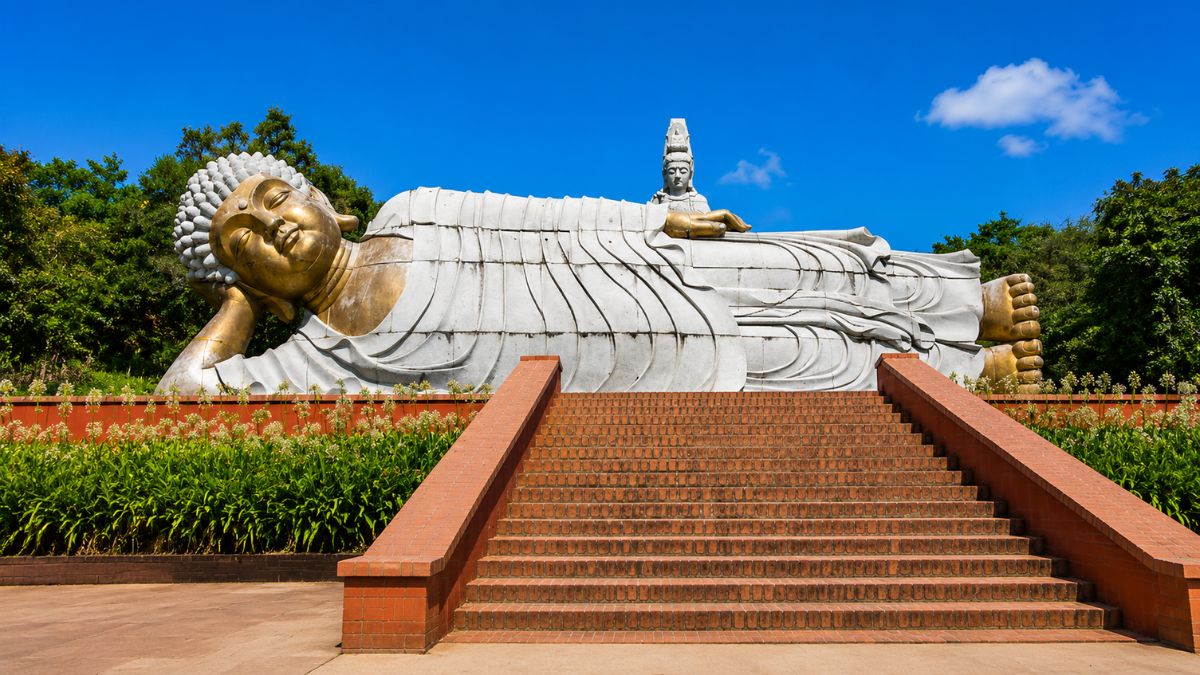 Uma gigantesca estátua dourada e branca do Buda Reclinado posicionada no topo de escadarias de tijolos vermelhos sob céu azul.