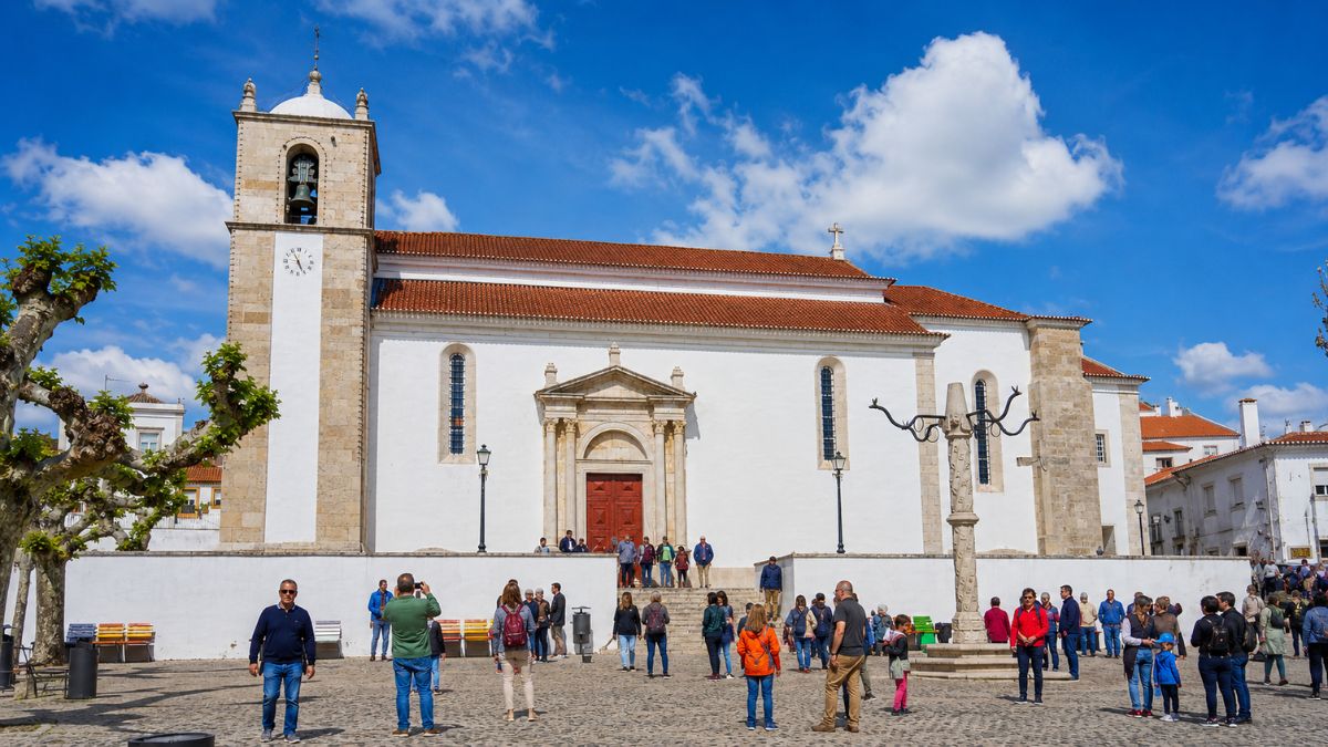 Fachada lateral branca da Igreja Matriz de Azambuja com portal de estilo serliano, torre sineira e cruzeiro de pedra em praça pública com visitantes.