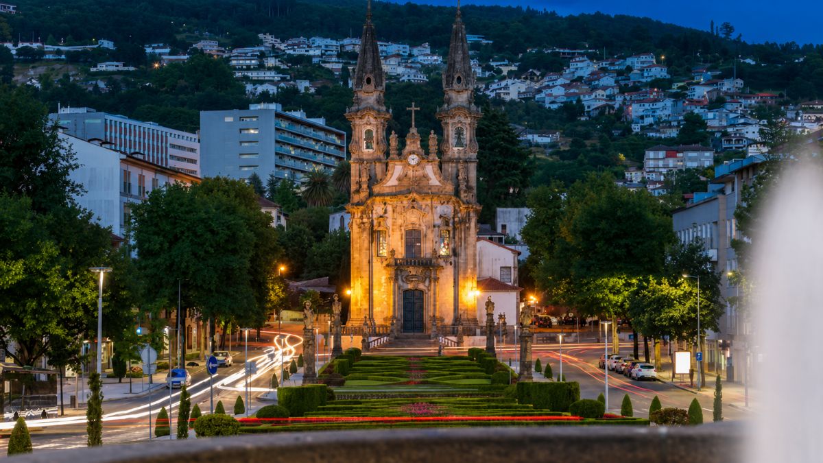 Vista panorâmica noturna do Largo da República do Brasil em Guimarães, Portugal, destacando a imponente Igreja de Nossa Senhora da Consolação iluminada ao fundo, com seus jardins simétricos em primeiro plano e rastros de luzes de carros em movimento nas laterais.