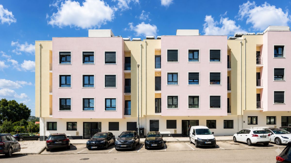 Fachada de prédio residencial moderno com tons rosa e amarelo e estacionamento com carros na freguesia central de Arruda dos Vinhos.