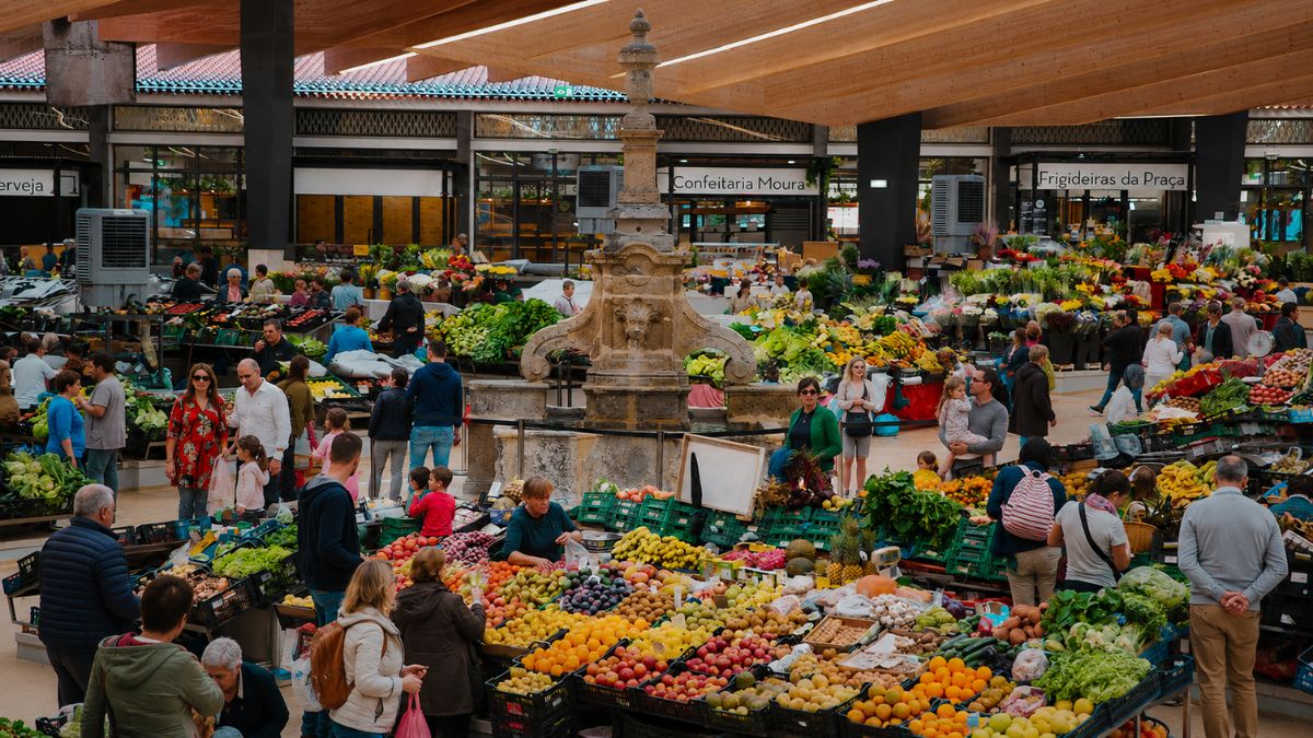 Vista aérea de mercado municipal movimentado em Braga com bancas cheias de frutas coloridas, vegetais frescos e pessoas fazendo compras sob uma estrutura moderna.