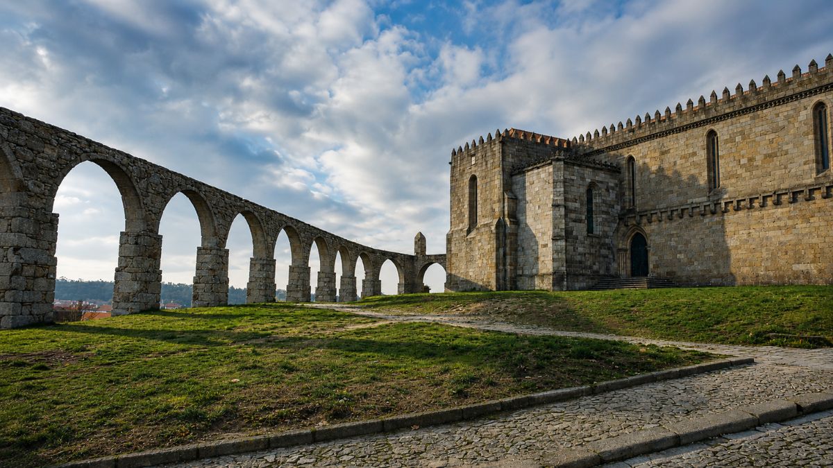 Uma estrutura arqueada de pedra à esquerda liga-se a uma imponente igreja gótica de granito à direita sobre uma colina relvada.