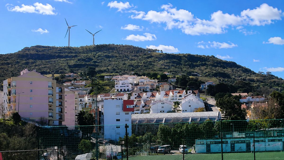 Vista panorâmica de Arruda dos Vinhos mostrando casas brancas e edifícios residenciais integrados às colinas verdes com aerogeradores de energia eólica sob céu azul.