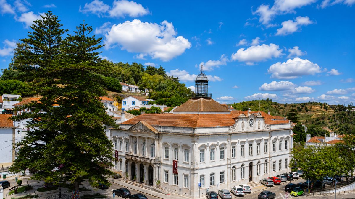 Vista aérea do edifício branco dos Paços do Concelho de Alenquer com telhado de telhas cerâmicas e uma torre central metálica sob céu azul com nuvens.