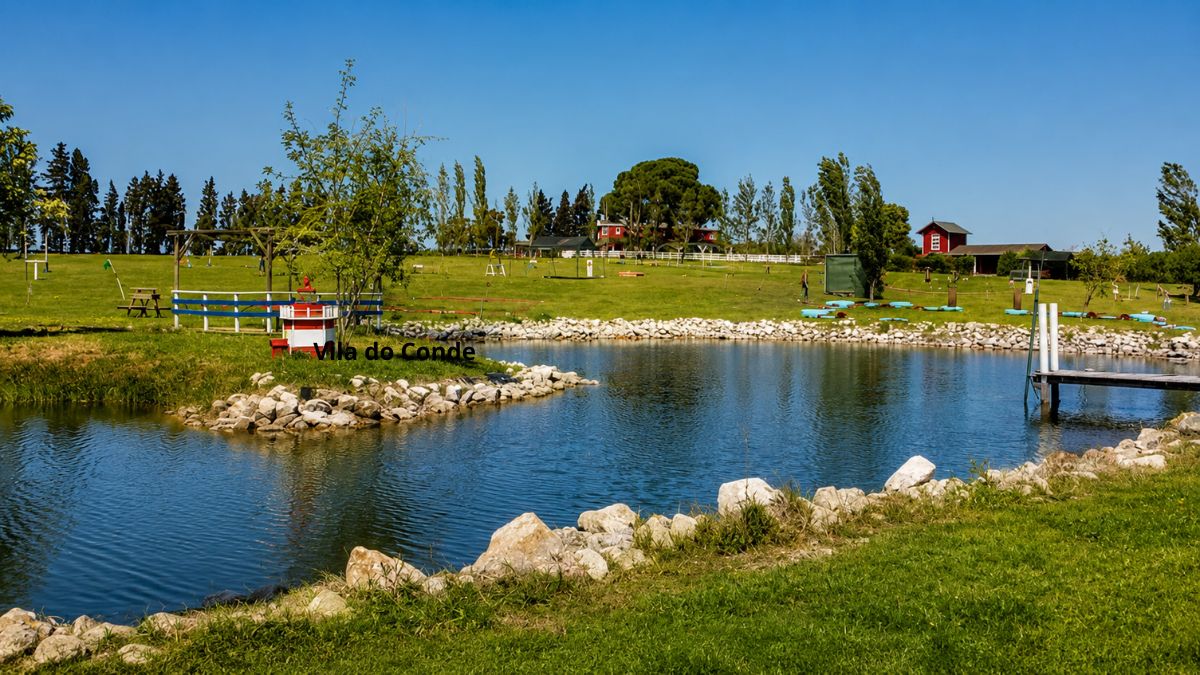 Um lago sereno cercado por pedras brancas e gramado verde, exibindo uma réplica de farol vermelho e branco na margem esquerda, uma pequena ponte de madeira à direita e construções rurais ao fundo sob um céu azul limpo.