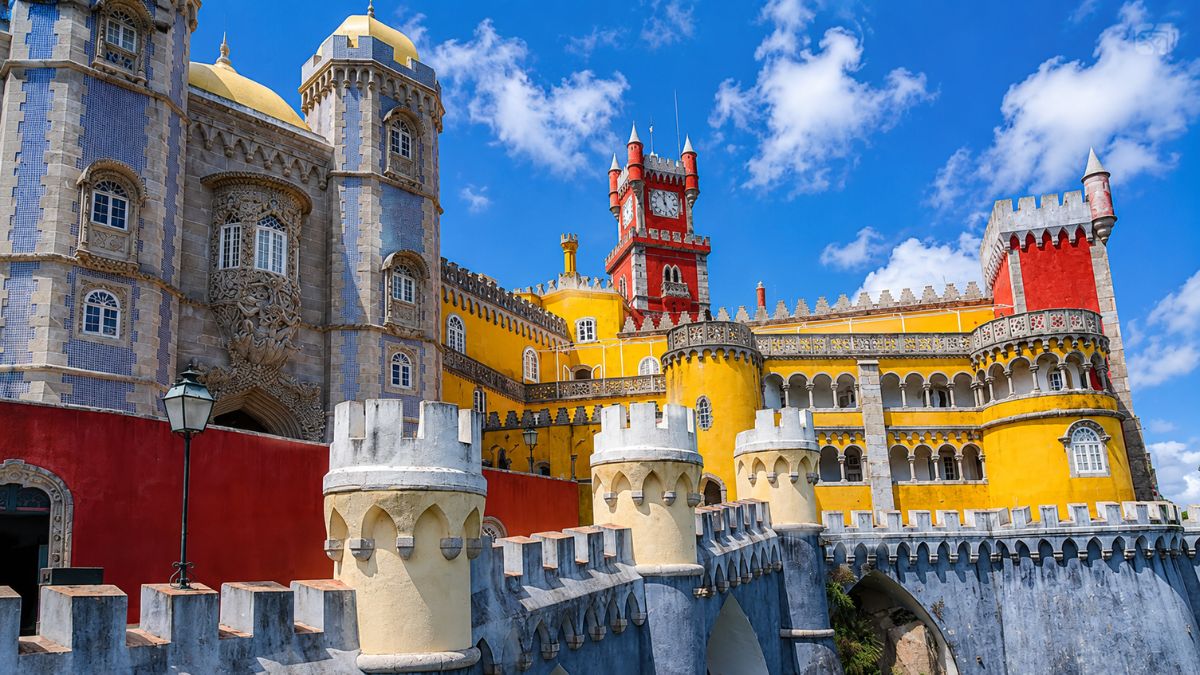 Vista angular das torres coloridas em tons de amarelo, vermelho e azul do Palácio da Pena sob céu azul com nuvens.