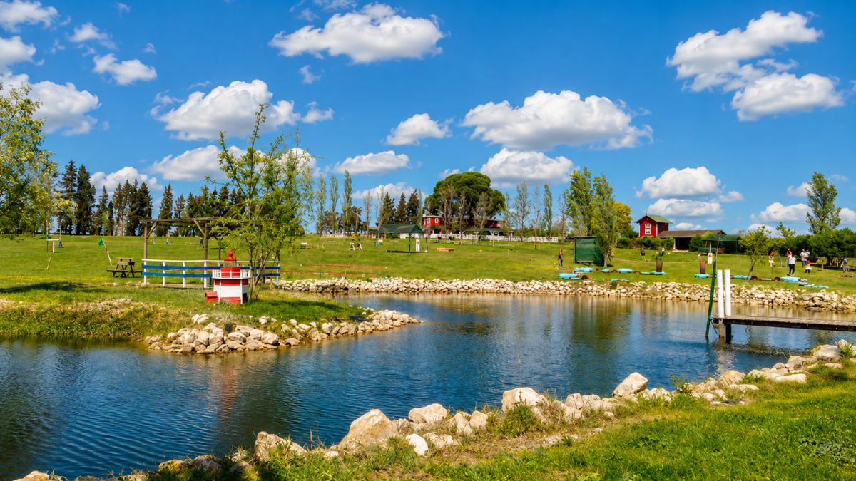 Vista panorâmica de um parque rural em Alenquer com lago artificial de águas azuis, cercado por pedras, gramado verde, árvores, pequenas casas vermelhas e céu ensolarado com nuvens.