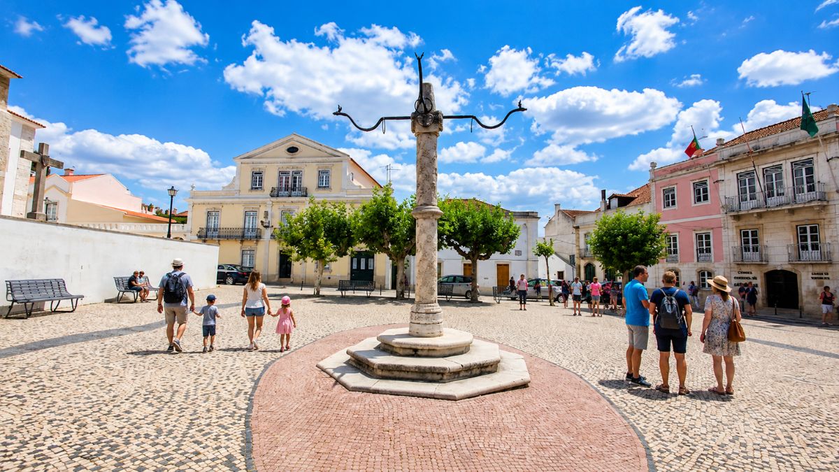 Turistas caminhando pela praça de paralelepípedos ao redor do monumento histórico do Pelourinho de Azambuja, com prédios antigos e céu azul ao fundo.
