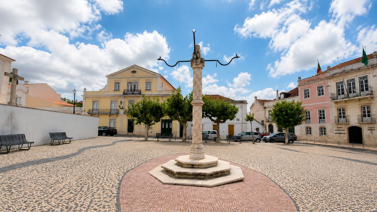 Fotografia da praça central de Manique do Intendente com o pelourinho histórico em destaque sobre calçada portuguesa e edifícios antigos ao fundo.