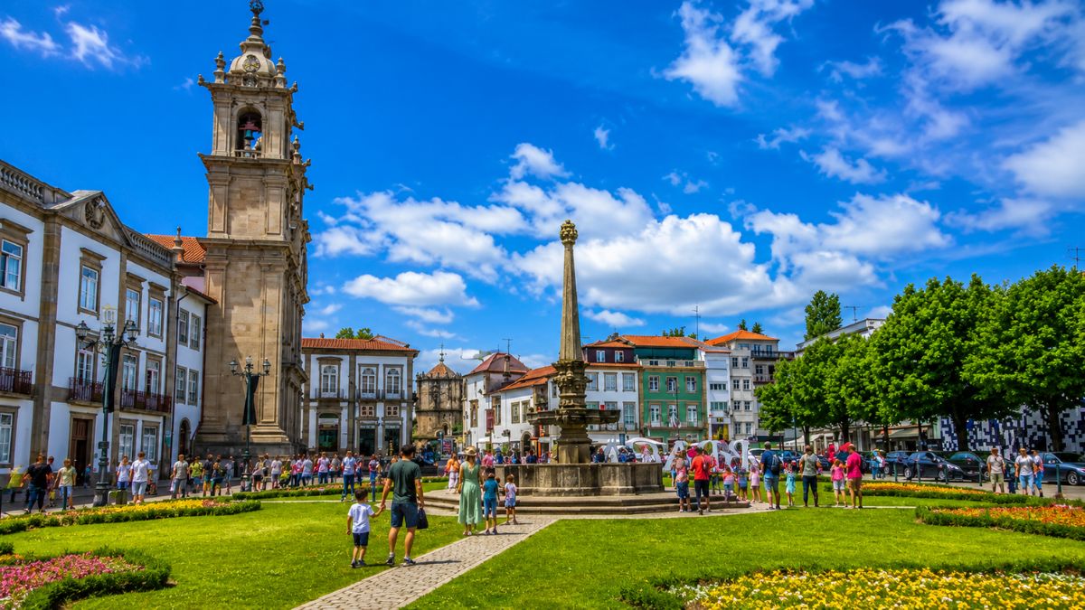 Vista da Praça da República em Braga com gramado verde, canteiros de flores, chafariz central, edifícios históricos e pedestres sob céu azul.