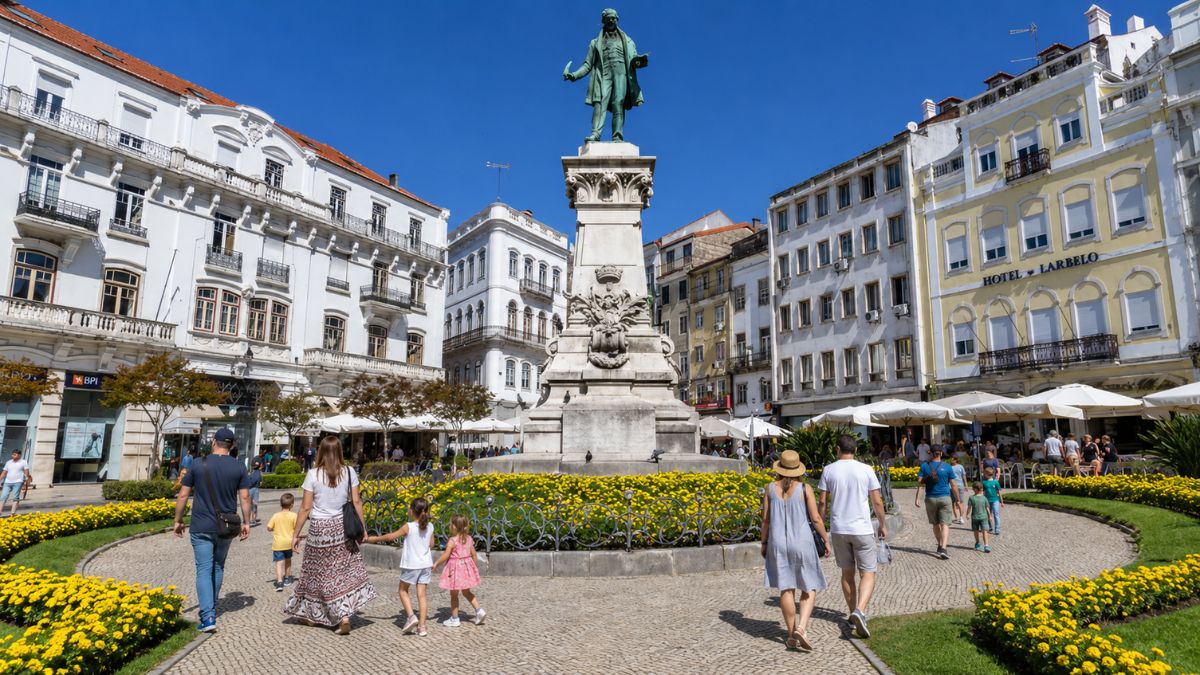 Vista da Praça do Comércio em Coimbra Portugal mostrando pessoas caminhando em dia ensolarado ao redor do monumento central com flores amarelas e edifícios históricos ao fundo incluindo o Hotel Larbelo.