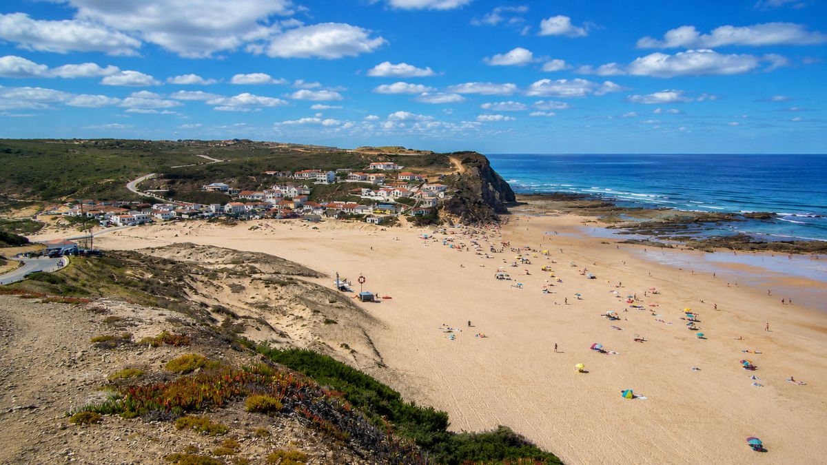 Vista panorâmica da Praia de Monte Clérigo no Algarve com sua ampla faixa de areia, falésias costeiras, mar azul e vilarejo ao fundo sob céu com nuvens.