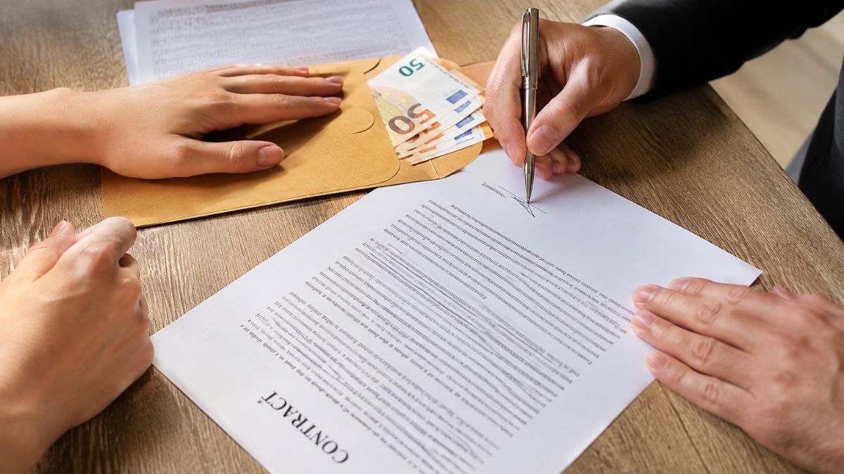 Mãos de duas pessoas assinando um contrato com caneta e um envelope com notas de 50 euros sobre uma mesa de madeira.