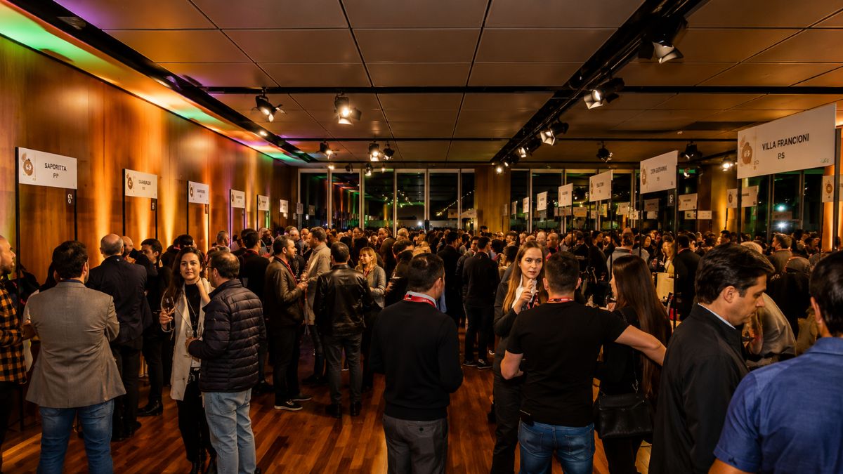 Fotografia de um amplo salão de degustação lotado durante o evento Vinhos de Portugal com pessoas conversando e taças na mão.