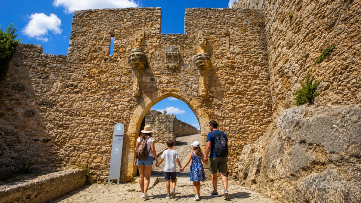 Família caminhando de mãos dadas através do portal de pedra de uma fortificação histórica de arquitetura medieval e militar.