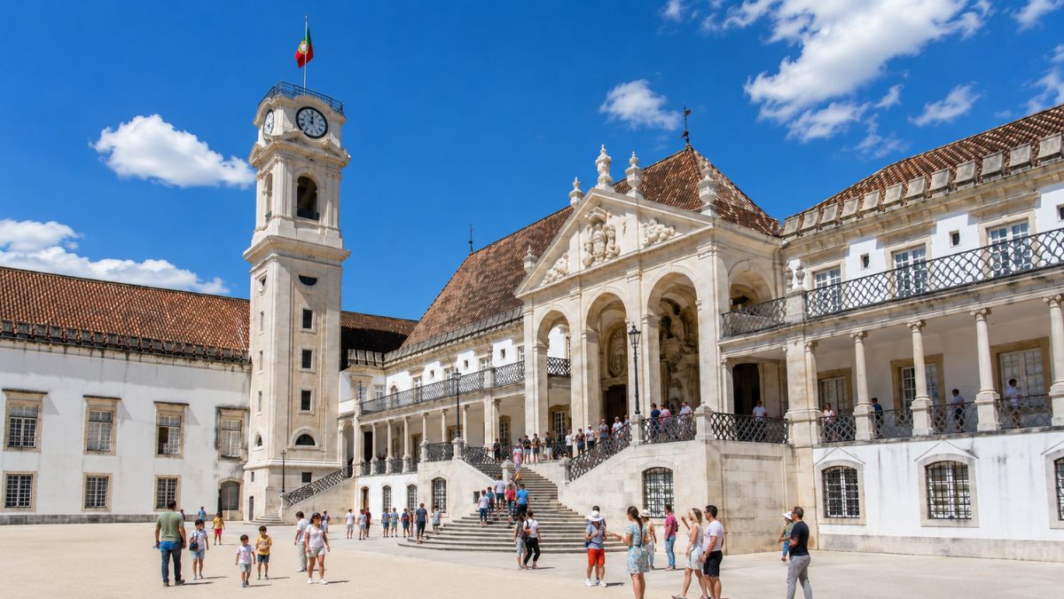 Fachada do pátio histórico da Universidade de Coimbra em Portugal com turistas circulando e a famosa torre do relógio sob o céu azul.