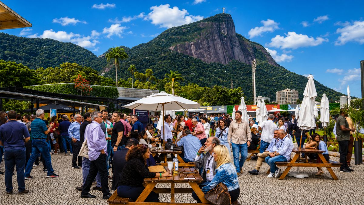 Pessoas reunidas em mesas ao ar livre no evento Vinhos de Portugal no Jockey Club na Gávea com vista para o Cristo Redentor e o Morro do Corcovado no Rio de Janeiro.