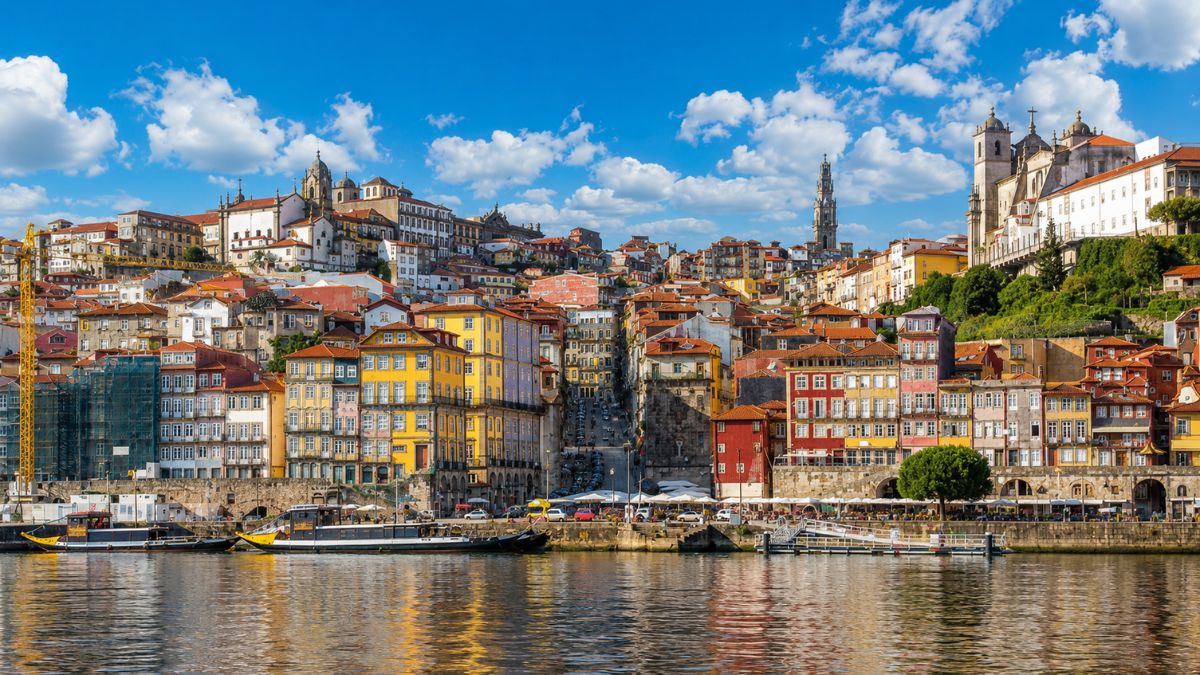 Vista panorâmica do Cais da Ribeira no Porto, Portugal, mostrando as fachadas coloridas dos edifícios medievais à beira do Rio Douro, barcos rabelos ancorados nas águas calmas e a icônica Torre dos Clérigos ao fundo sob um céu azul com nuvens brancas.