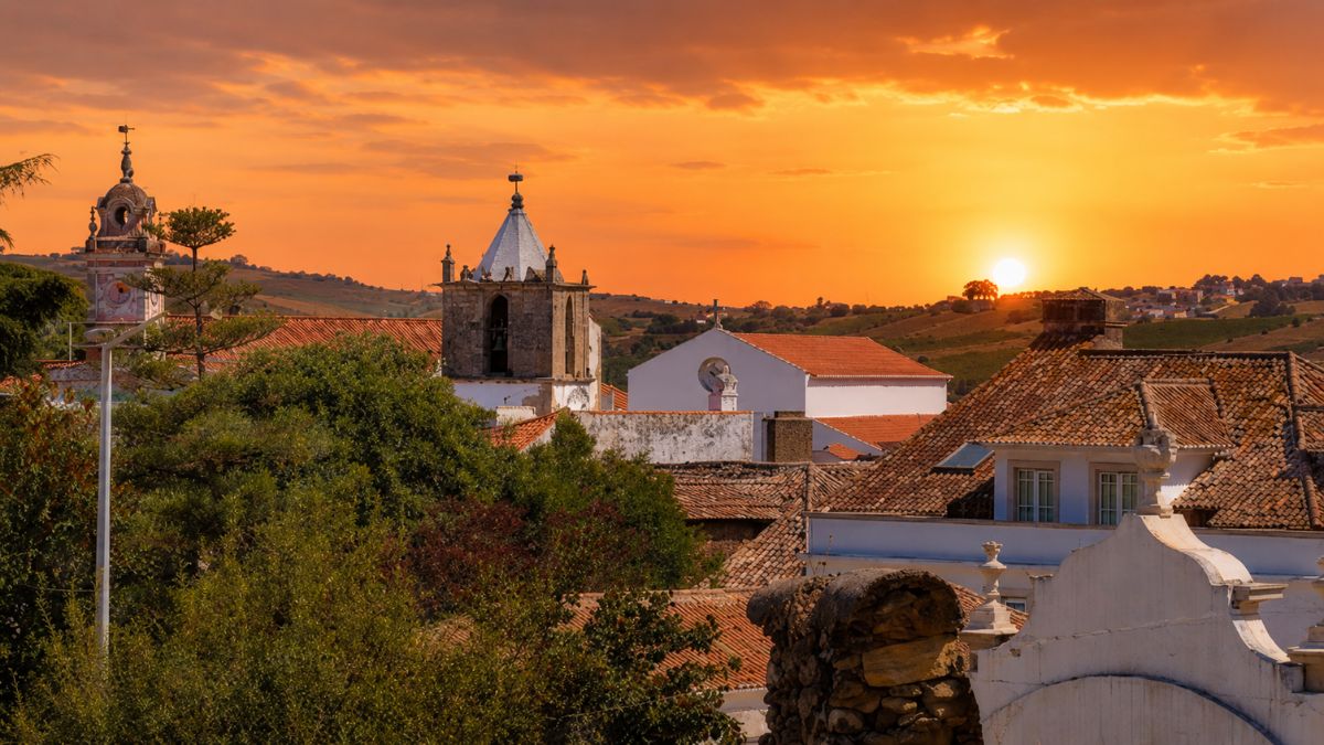 Vista aérea dos telhados antigos e torres das igrejas de Arruda dos Vinhos sob um céu alaranjado durante o pôr do sol.