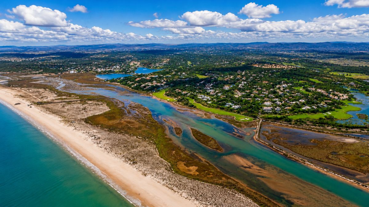 Fotografia aérea mostrando a Praia da Quinta do Lago à esquerda com mar azul e areia clara, dividida por canais de água da Ria Formosa que margeiam campos de golfe verdes e mansões luxuosas ao fundo sob céu com nuvens brancas.
