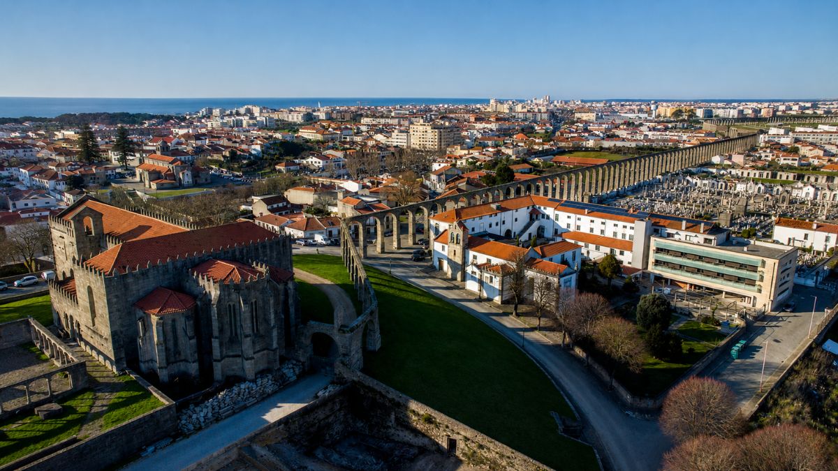 Vista aérea panorâmica mostrando a imponente estrutura de pedra do Mosteiro de Santa Clara em primeiro plano e o extenso Aqueduto histórico cortando a paisagem urbana da cidade de Vila do Conde com o oceano ao fundo.
