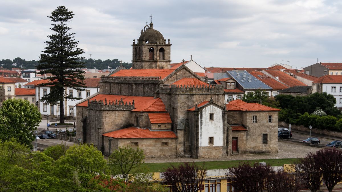 Uma grande igreja antiga construída em blocos de pedra escura com vários telhados de telha laranja e uma torre sineira abobadada ao centro.