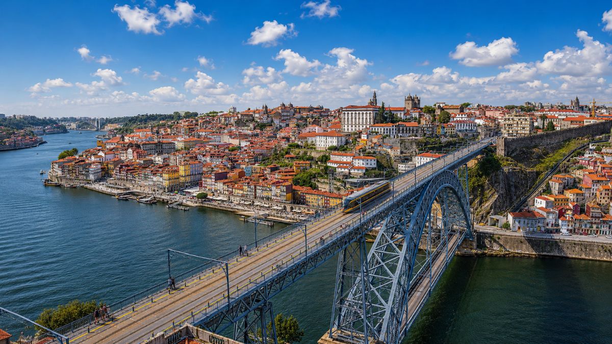 Fotografia diurna da Ponte Luís I cruzando o Rio Douro na cidade do Porto, mostrando um trem do metrô sobre a estrutura metálica e o casario colorido da Ribeira ao fundo sob um céu azul com nuvens.