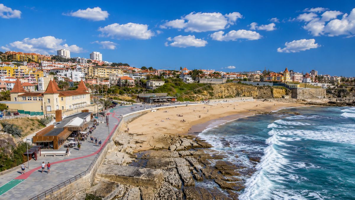 Fotografia colorida da Praia do Tamariz com faixa de areia dourada banhada pelo Oceano Atlântico e a encosta residencial do Monte Estoril ao fundo sob céu azul com nuvens.